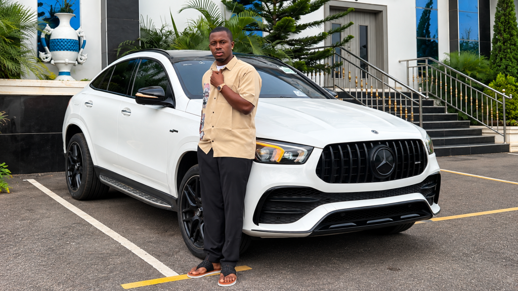 Prince Dumebi Micheal standing beside a white Mercedes-Benz SUV outdoors.