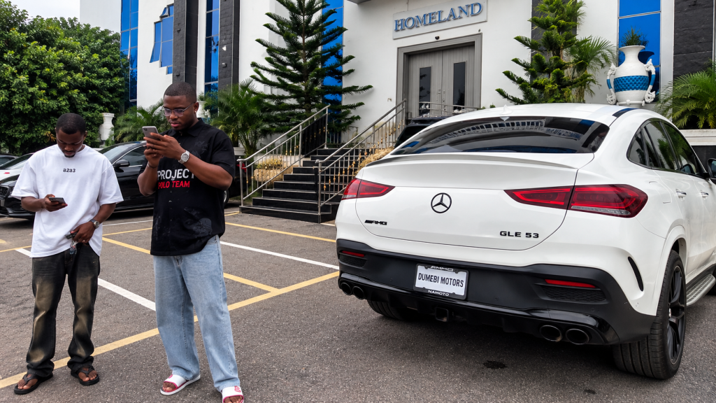 Prince Dumebi Micheal standing near a white Mercedes-AMG GLE 53 displaying Dumebi Motors branding.
