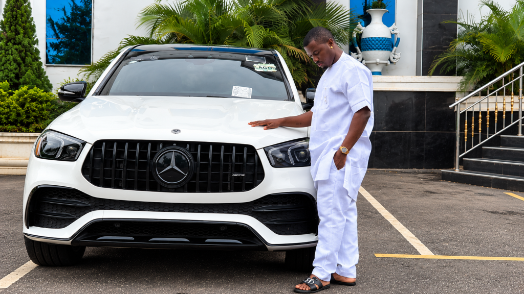 Prince Dumebi Micheal standing beside a white Mercedes-Benz SUV with one hand resting on the hood.